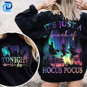 Peut inclure: Sweat-shirt noir avec un motif coloré représentant des silhouettes de sorcières volant sur des balais devant une aurore boréale vibrante. Le texte dit "It's Just a Bunch of Hocus Pocus" et "Tonight We Fly".