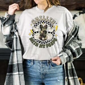 May include: A light gray sweatshirt with a graphic that says "Chillin' With My Peeps." The graphic features three cartoon bunnies wearing sunglasses, with one in camouflage. The design is surrounded by a black and white checkered circle, lightning bolts, and stars.