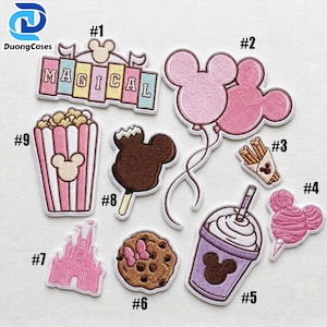 May include: A collection of nine embroidered patches featuring various designs including a sign reading MAGICAL, pink mouse-shaped balloons, churros, popcorn, a chocolate-covered treat, a castle, a cookie, a drink, and cotton candy.