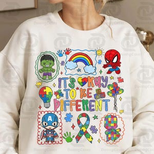 May include: White sweatshirt with a colorful graphic design featuring the text "IT'S OKAY TO BE DIFFERENT" and illustrations of superheroes, a rainbow, puzzle pieces, and a ribbon.
