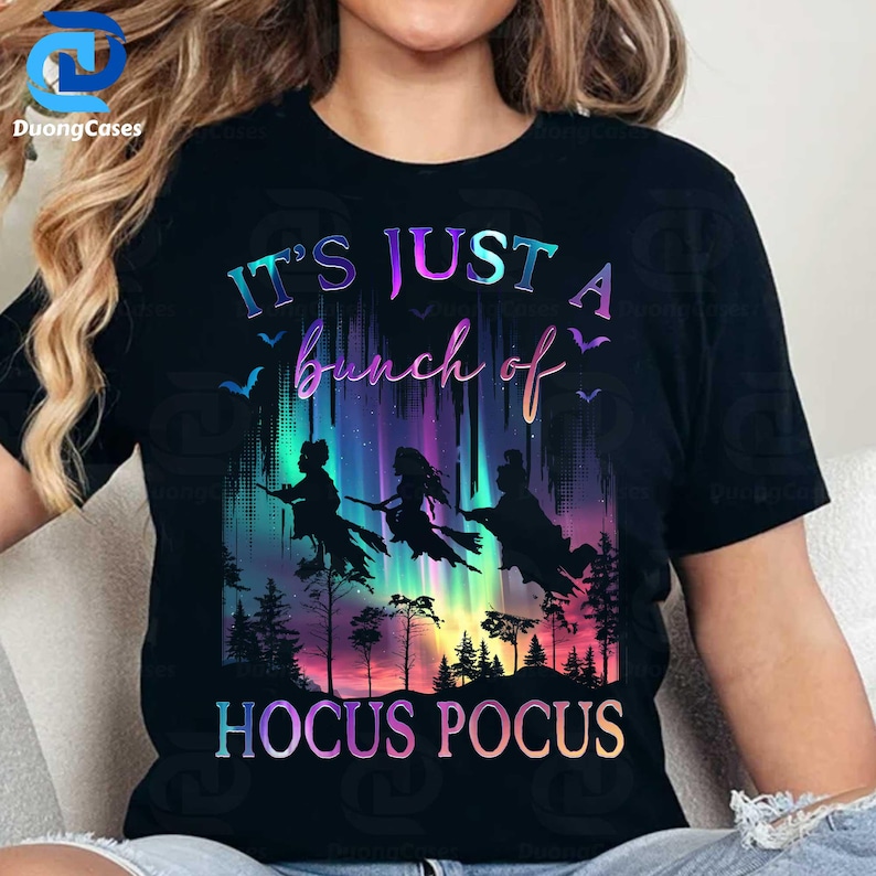 May include: Black t-shirt with the text "IT'S JUST A bunch of HOCUS POCUS" in a colorful, gradient font. The graphic features silhouettes of three witches flying on broomsticks against a vibrant, colorful background with trees and bats.