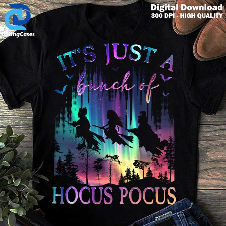May include: Black t-shirt with a colorful graphic featuring three witch silhouettes flying in front of a vibrant aurora borealis. The text reads "IT'S JUST A bunch of HOCUS POCUS" in a gradient font, perfect for Halloween.