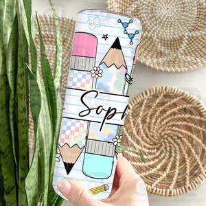 May include: A white tumbler with a pencil and eraser design, featuring the name "Sop" in black script. The design includes a pencil, eraser, flowers, and molecular structures. The tumbler is held by a person.
