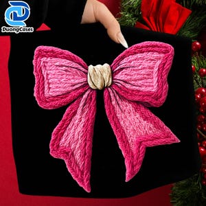 May include: Black accessory featuring a large, embroidered pink bow. The bow has a textured appearance with varying shades of pink and a cream-colored center knot. The logo "DuongCases" is visible in the upper left corner.