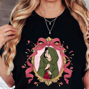 Puede incluir: Camiseta negra con una ilustraci&oacute;n de dibujos animados de un personaje con una capa con capucha verde dentro de un marco rosa y dorado. El marco est&aacute; adornado con un lazo rosa y detalles florales. El personaje sostiene una bolsa roja.