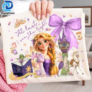 May include: A cream-colored tote bag featuring a princess illustration with long blonde hair, wearing a purple dress. The bag has a purple bow, a castle, and the text "At last I see the light".