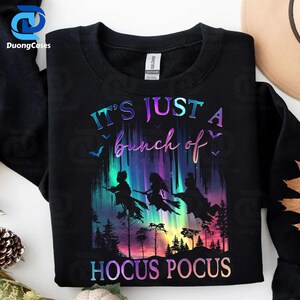 May include: Black sweatshirt with the text "IT'S JUST a bunch of HOCUS POCUS" in a colorful, holographic font. The design features silhouettes of three witches flying on brooms against a vibrant, colorful background.