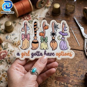 May include: An embroidered patch featuring five stylized broomsticks with Halloween motifs including ghosts, pumpkins, a spider web, and a bat. The text "a girl gotta have options" is stitched in orange and purple letters at the bottom of the patch.