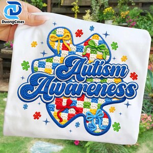 May include: White t-shirt with a colourful puzzle piece design and the words "Autism Awareness" in blue. The puzzle piece has a checkered pattern with bows and puzzle piece accents in various colours.