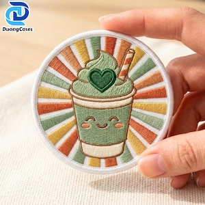 May include: A circular embroidered patch featuring a green cup with a smiling face, topped with whipped cream and a heart shape. The background consists of radiating stripes in shades of orange, yellow, and green, all enclosed by a white border.