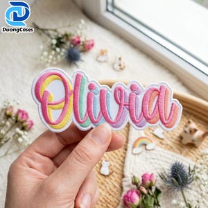 Könnte beinhalten: Ein personalisierter Namensaufnäher mit dem Namen Olivia in Schreibschrift. Die Buchstaben sind mit einem mehrfarbigen Verlaufseffekt gestickt, der von Rosa über Gelb und Hellblau bis hin zu Violett reicht, umrandet von einer weißen Linie.