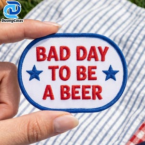 Może przedstawiać: Owalna haftowana naszywka z białym tłem i niebieską obwódką. Naszywka zawiera wyhaftowany czerwoną nicią napis "BAD DAY TO BE A BEER" ułożony w trzech liniach, z niebieską wyhaftowaną gwiazdką po obu stronach środkowej linii.
