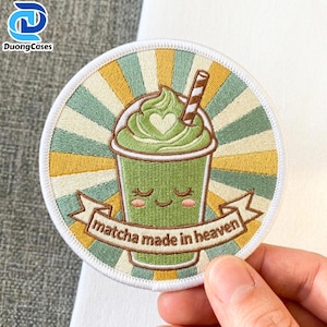 May include: A circular embroidered patch featuring a green matcha drink with a heart-shaped foam top and a striped straw. The background has yellow and green sunburst rays. A brown banner at the bottom reads "matcha made in heaven".