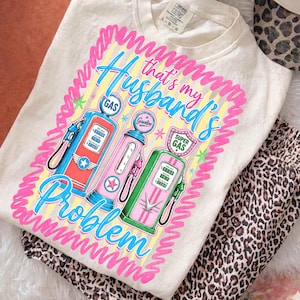 Pode incluir: Camiseta creme com um design gráfico com três bombas de gasolina vintage em vermelho, rosa e verde, com o texto "That's my Husband's Problem" em azul e rosa. A camisa é dobrada com calças com estampa de leopardo.