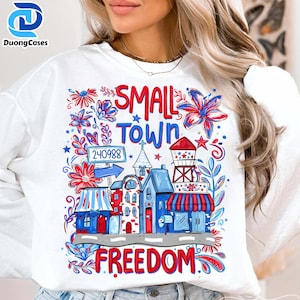 Könnte beinhalten: Weißes Sweatshirt mit einem farbenfrohen Grafikdesign, das eine Kleinstadt zeigt. Das Design enthält die Worte "SMALL TOWN FREEDOM" und eine Stadtillustration mit Gebäuden, Blumen und einem Wasserturm. Die Nummer "240988" ist ebenfalls enthalten.