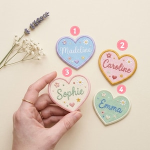 May include: Four heart-shaped embroidered patches with names Madeline, Caroline, Sophie, and Emma. Each patch features floral and star motifs in pastel colors with a stitched border. The patches are numbered one through four in pink circles.