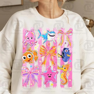 May include: White sweatshirt featuring colorful cartoon sea creatures and bows on a pink striped background. The design includes an octopus, shark, fish, seahorse, starfish, and bows in various colors. The sweatshirt has a crew neck.