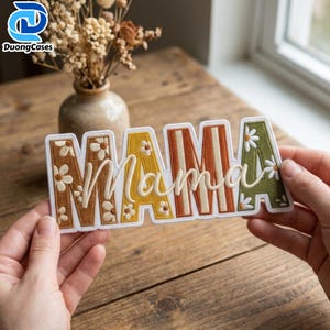 May include: A rectangular embroidered patch featuring the word MAMA in block letters with a white outline. The letters are filled with a floral pattern in shades of brown, yellow, orange, and green, with the word mama in white cursive script.