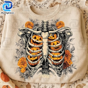 May include: Beige sweatshirt featuring a Halloween-themed design. The graphic depicts a rib cage filled with carved jack-o'-lanterns, surrounded by floral elements. The text "DuongCases" is visible in the upper left corner.