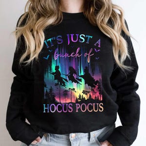 May include: Black sweatshirt with a colorful graphic featuring three witches on broomsticks flying in front of a vibrant aurora borealis. The text "IT'S JUST A bunch of HOCUS POCUS" is displayed in a gradient font.