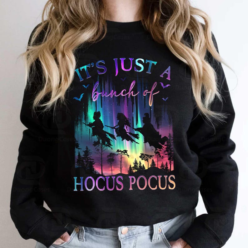 May include: Black sweatshirt with a colorful graphic featuring three witches on broomsticks flying in front of a vibrant aurora borealis. The text "IT'S JUST A bunch of HOCUS POCUS" is displayed in a gradient font.