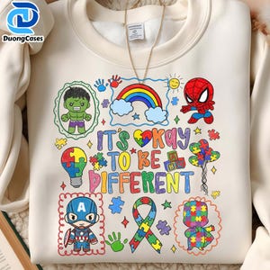 May include: Cream-colored sweatshirt with a colorful graphic design. The design includes cartoon characters, a rainbow, puzzle pieces, and the text "IT'S OKAY TO BE DIFFERENT". The sweatshirt has a crew neck and long sleeves.