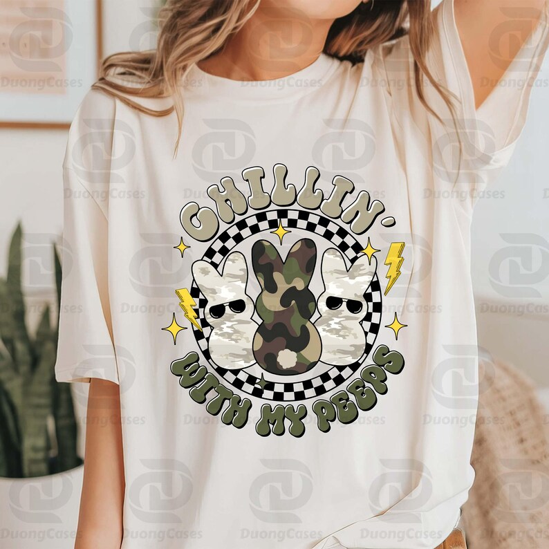 May include: Cream-colored t-shirt featuring a graphic with the text "CHILLIN' WITH MY PEEPS." The design includes three cartoon bunnies wearing sunglasses, one in camouflage, set against a black and white checkered background with yellow lightning bolts and stars.