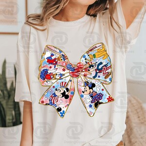 K&ouml;nnte beinhalten: Ein wei&szlig;es T-Shirt mit einem gro&szlig;en, bunten Schleifendesign. Die Schleife besteht aus verschiedenen Disney-Charakteren, darunter Mickey Mouse, Minnie Mouse, Donald Duck und Goofy. Die Charaktere tragen rot-wei&szlig;-blaue Kleidung, und die Schleife ist mit Sternen und Streifen verziert.