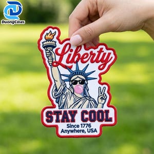 Pode incluir: Um emblema bordado com a Estátua da Liberdade usando óculos de sol pretos e soprando uma bolha de chiclete rosa. O texto diz Liberty, STAY COOL, Since 1776 e Anywhere, USA. O design utiliza fios nas cores vermelha, branca, azul e rosa.
