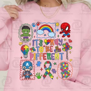 May include: Pink sweatshirt with colorful graphic featuring cartoon characters, a rainbow, and the text "IT'S OKAY TO BE DIFFERENT." The design includes puzzle pieces and a ribbon.
