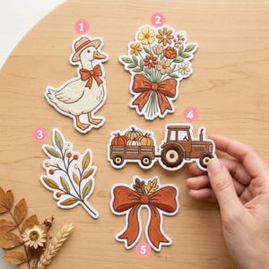 May include: A set of five die-cut stickers featuring autumn-themed illustrations: a goose wearing a hat and bow, a bouquet of fall flowers with a bow, a leafy branch with berries, a tractor pulling a wagon of pumpkins, and a large orange bow.