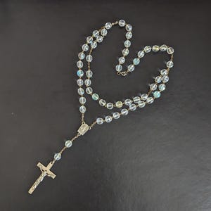 Puede incluir: Un rosario con cuentas de vidrio facetado, transparentes e iridiscentes, unidas por una fina cadena de metal dorado. El rosario incluye una pieza central rectangular y un crucifijo de metal dorado con la figura de Jesús en el extremo inferior.