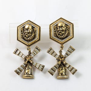 Puede incluir: Un par de pendientes de metal dorado con una pieza superior hexagonal que presenta un retrato en relieve de una persona con cuello de lechuguilla. Un dije de molino de viento con aspas texturadas cuelga de cada pieza hexagonal.