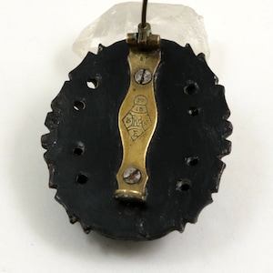 May include: The back of a black oval brooch with a gold pin and clasp. The brooch has a decorative edge and a series of holes around the perimeter. The pin has the text "5 K 18 E" on it.