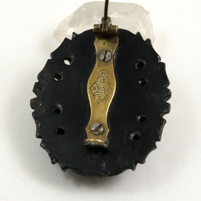 May include: The back of a black oval brooch with a gold pin and clasp. The brooch has a decorative edge and a series of holes around the perimeter. The pin has the text "5 K 18 E" on it.