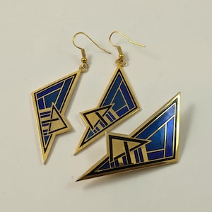 Puede incluir: Un conjunto de dos pendientes de metal dorado y un broche a juego, cada uno con un diseño geométrico con incrustaciones de esmalte azul profundo y contornos negros. Los pendientes cuelgan de ganchos dorados y el broche tiene forma angular.