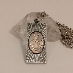 May include: Silver pendant necklace with a rectangular charm featuring a detailed image of the Virgin Mary holding a baby Jesus. The charm has a textured, sunburst design around the image, which is in rose gold.