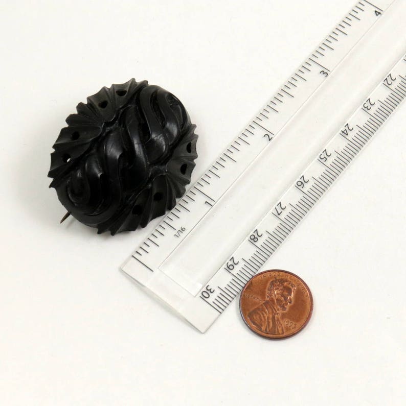 May include: A black carved jet brooch with a detailed design. The brooch is approximately 1 inch in diameter and is shown next to a ruler and a penny for scale.