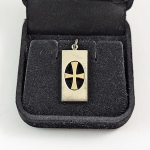 May include: A silver pendant with a gold cross design inside a rectangular frame. The pendant is displayed in a black jewelry box with a velvet lining. The cross is centered within an oval shape.
