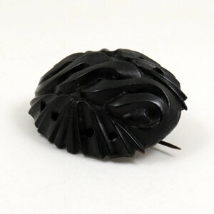 May include: A black carved jet brooch with a pin on the back. The brooch has a swirling design and is oval shaped.