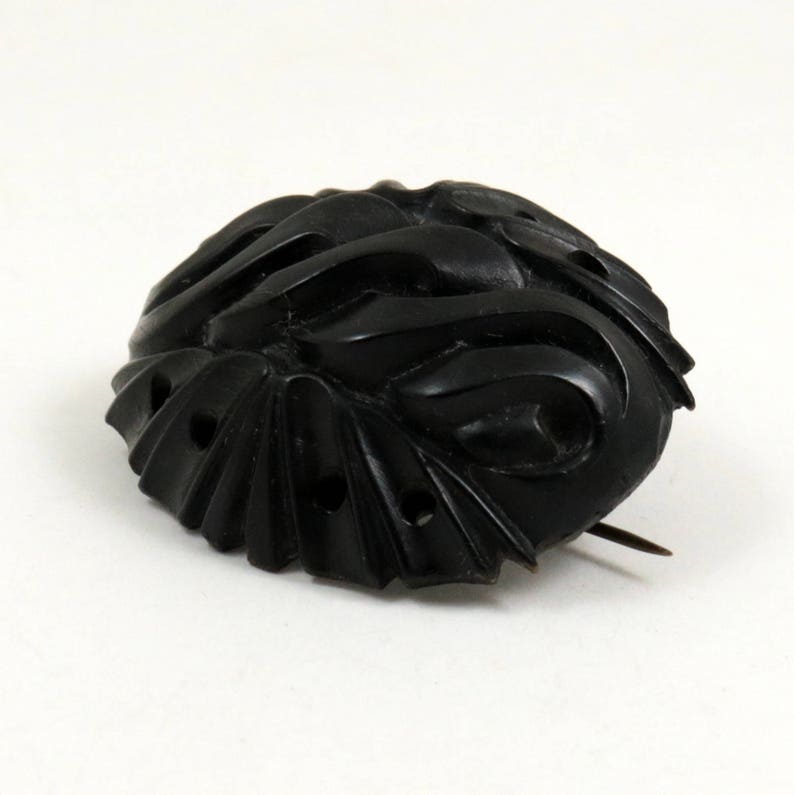 May include: A black carved jet brooch with a pin on the back. The brooch has a swirling design and is oval shaped.