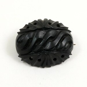 May include: A black carved jet brooch with a swirling pattern. The brooch is oval shaped and has a pin on the back.