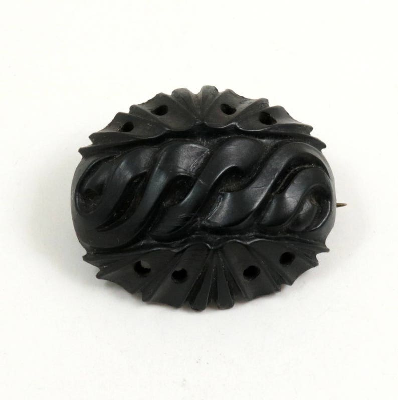 May include: A black carved jet brooch with a swirling pattern. The brooch is oval shaped and has a pin on the back.
