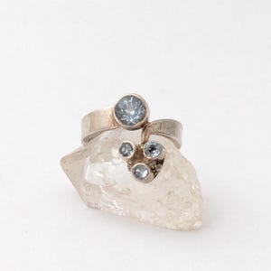 Könnte beinhalten: Ein Silberring mit einem großen, runden, hellblauen Edelstein und drei kleineren hellblauen Edelsteinen. Der Ring liegt auf einer klaren, kristallinen Oberfläche.