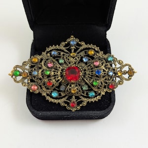 May include: Ornate vintage brooch with a filigree design in antique gold. The brooch features a large red center stone and is adorned with an array of colorful gemstones in shades of blue, green, yellow, pink, and orange. The brooch is displayed in a black velvet box.
