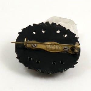 May include: The back of a black oval brooch with a gold pin and a gold inscription that reads "Made in England".