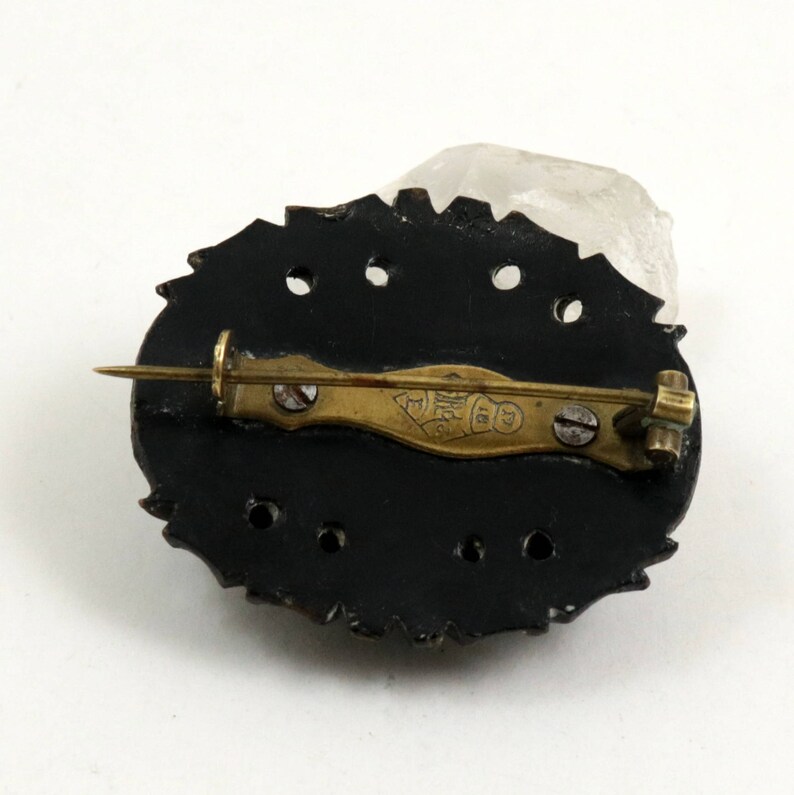 May include: The back of a black oval brooch with a gold pin and a gold inscription that reads "Made in England".
