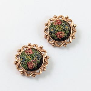 Puede incluir: Dos botones redondos con una incrustación central de tela con un patrón de bordado floral multicolor en tonos rosa, azul y verde sobre un fondo negro, rodeados por un marco decorativo de metal dorado con detalles en forma de hojas.