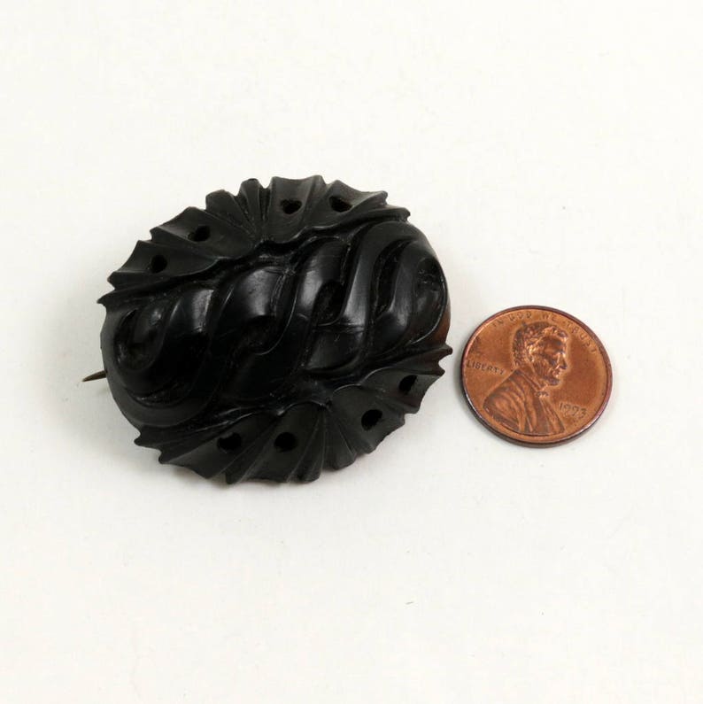 May include: A black carved jet brooch with an intricate design. The brooch is oval shaped and has a pin on the back.