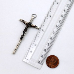 Puede incluir: Pendiente de cruz de plata con una figura de Jes&uacute;s de color marr&oacute;n oscuro. El pendiente mide aproximadamente 5 cm de largo y se muestra junto a una regla y una moneda de un centavo para comparar el tama&ntilde;o.